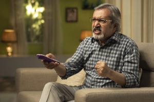 Mature man sitting on sofa angrily watching tv with remote in hand